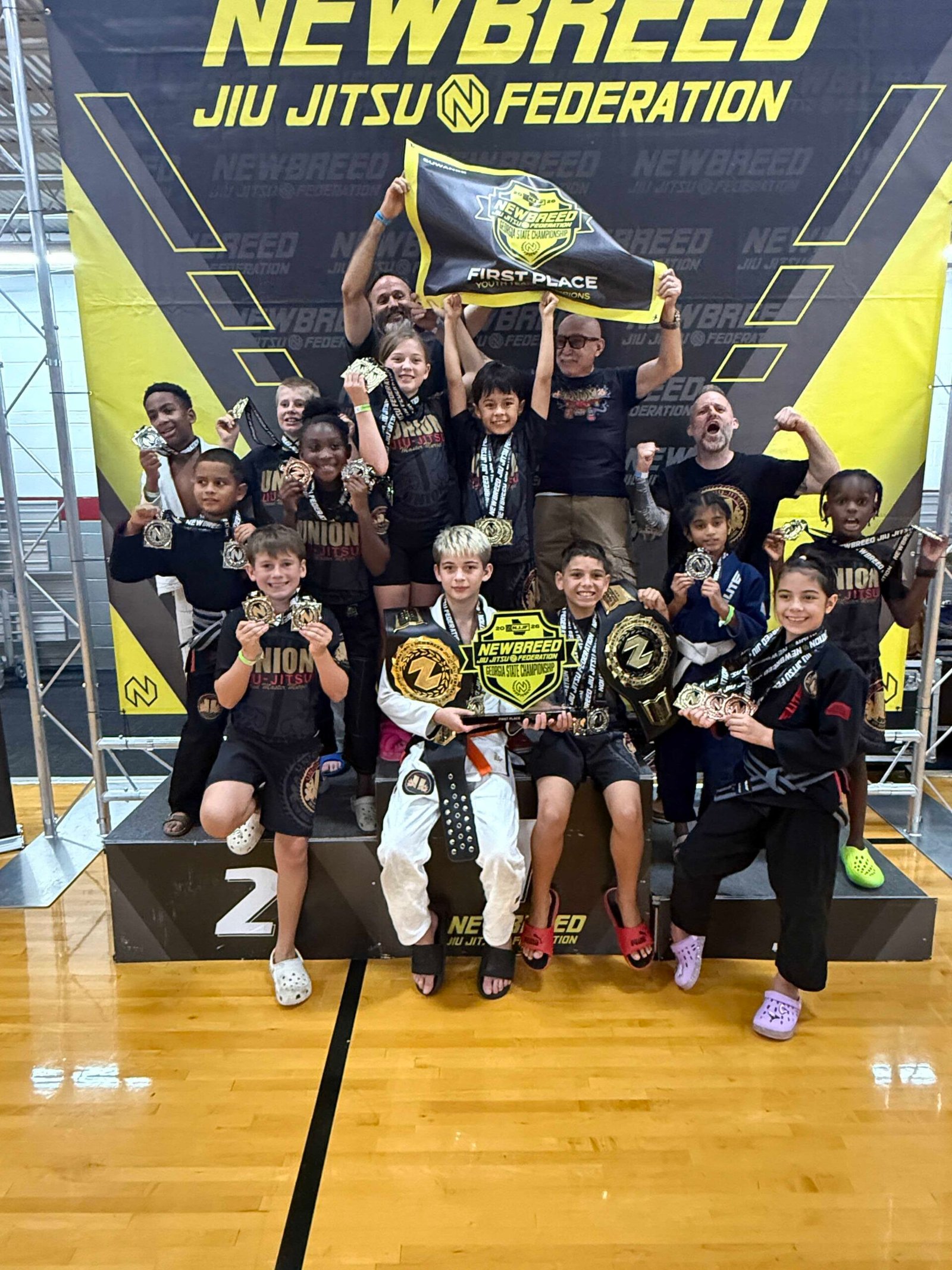 Union Team BJJ coaches and students with medals, championship belts, and trophy at a Newbreed Jiu Jitsu Federation competition
